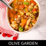 A steaming bowl of Olive Garden Pasta Fagioli soup rests elegantly on a white surface, accompanied by a spoon and nestled atop a red-striped cloth.