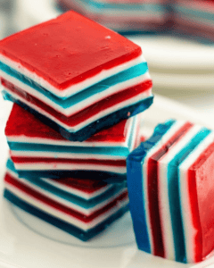 Three vibrant, layered Jello squares in red, white, and blue are stacked on a plate, with more similar squares in the background.