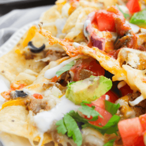 A delicious plate of loaded nachos is generously topped with melted cheese, creamy avocado, juicy tomatoes, spicy jalapeños, zesty onions, and a dollop of tangy sour cream for the ultimate snacking experience.