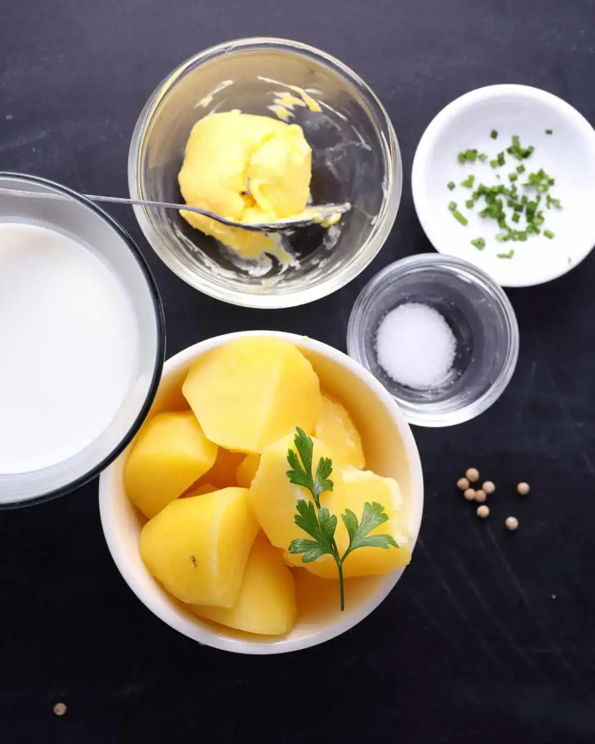 Bowl of peeled vegetables, cup of milk, small bowl of butter, dish of salt, peppercorns, and chopped herbs.